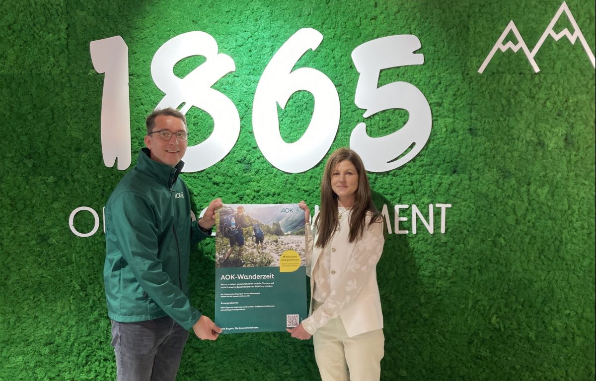 Stefan Rampp (links) von der AOK und Marktleiterin Melanie Palta (rechts) von 1865 Outdoor Equipment Kaufbeuren mit dem Plakat zur AOK-Wanderzeit 2025. Foto: AOK Bayern, Stefan Rampp